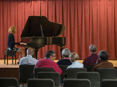 Six senior men and women sitting in Whitney Center's Cultural Arts Center, listening to a woman playing a grand piano