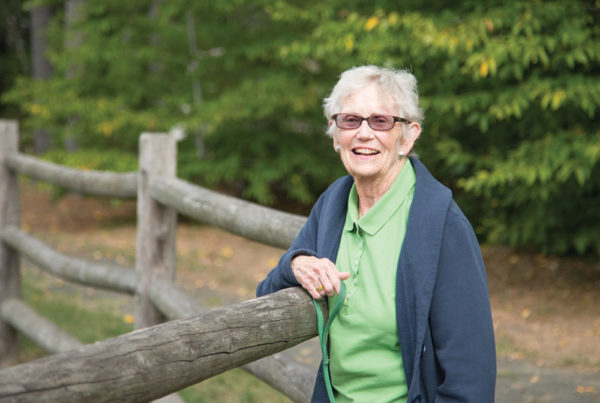 A smiling senior woman leans against a wooden railing on one of Whitney Center's on-campus walking trails