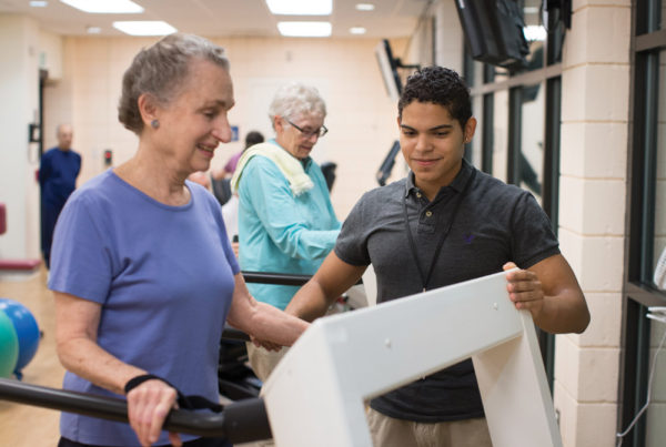 A young male staff member assists a smiling senior woman on the treadmill in the Whitney Center fitness center